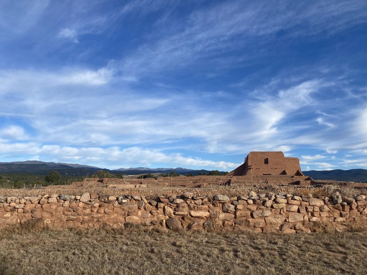 Ancient adobe structure under a blue sky with clouds and distant mountains.