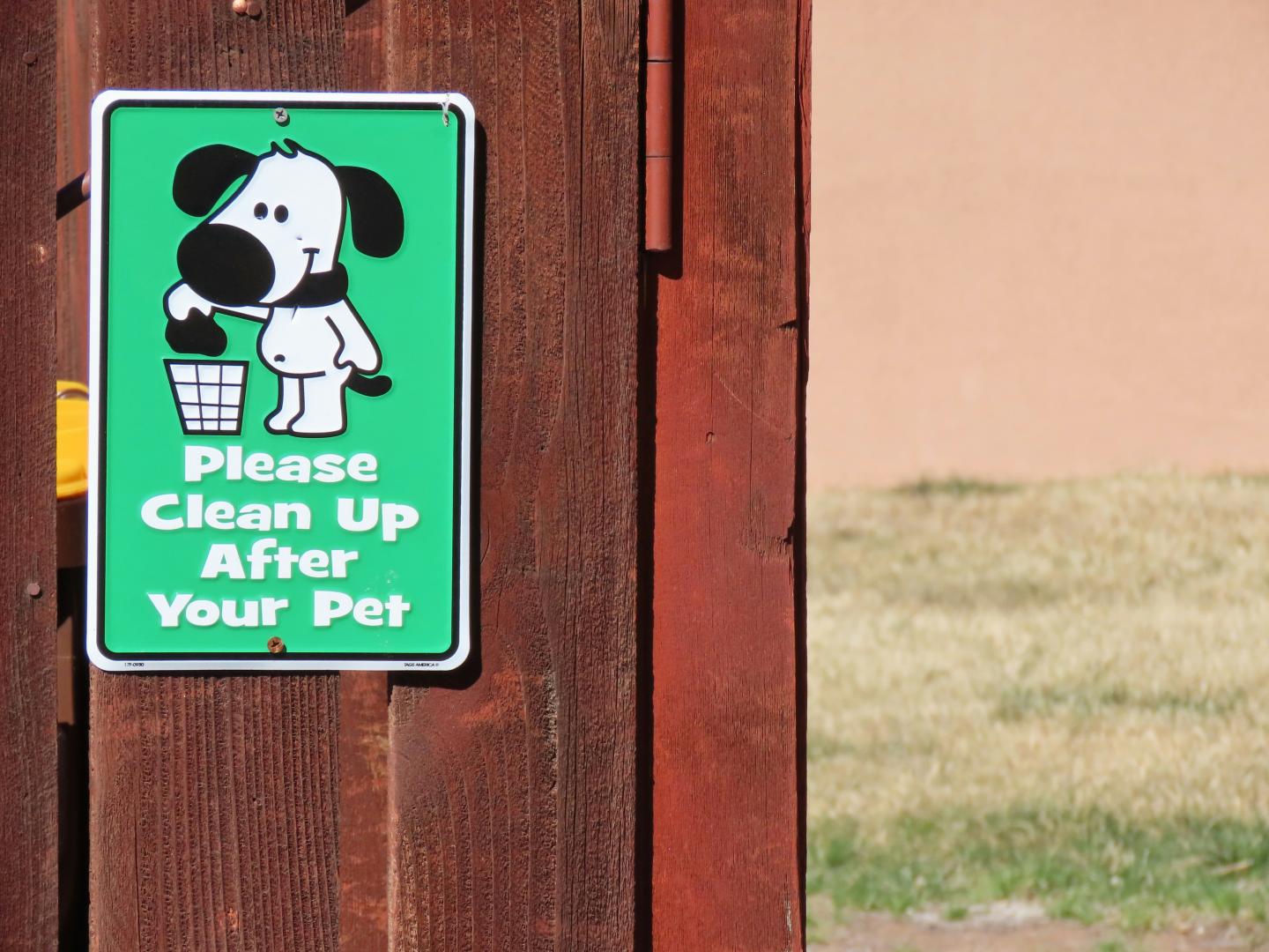 Green sign with cartoon dog, urging pet cleanup, on a brown wooden fence.